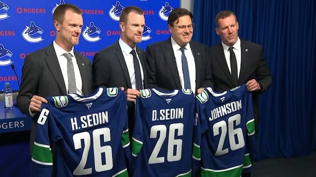 Four men in suits hold Vancouver Canucks jerseys at a press conference announcing front office appointments